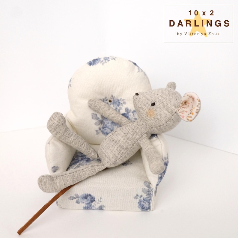 May include: A grey stuffed mouse toy with a pink-patterned ear rests on a small armchair. The armchair is upholstered in white fabric with blue floral patterns. A brown stick is in the foreground. The text "10 x 2 DARLINGS by Viktoriya Zhuk" is in the upper right corner.