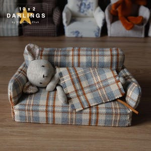 May include: A miniature sofa with a blue, cream, and brown plaid pattern, with a gray stuffed bunny. The sofa has a matching plaid blanket. The text "10 x 2 DARLINGS by Viktoriya Zhuk" is visible. Other miniature chairs are in the background.