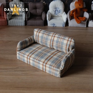May include: A miniature sofa with a blue, beige, and brown plaid pattern. The small couch has a rectangular shape with a backrest and armrests. The image also shows other miniature chairs in the background. Text on the image reads "10 x 2 DARLINGS by Viktoriya Zhuk".