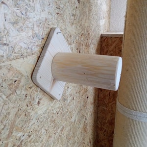 Peut inclure: Un accessoire en bois pour griffoir pour chat. L'accessoire est en bois non fini et se compose d'une base triangulaire avec un poteau cylindrique s'étendant vers l'extérieur. Il est monté sur un mur texturé brun clair.