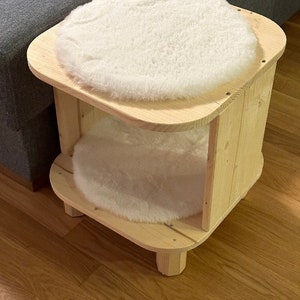 Könnte beinhalten: Ein helles Haustierbett aus Holz mit zwei Ebenen. Jede Ebene hat ein weiches, weißes, flauschiges Kissen. Das Bett hat ein schlichtes, rustikales Design mit abgerundeten Ecken und kurzen Beinen, geeignet für einen kleinen Hund oder eine Katze.