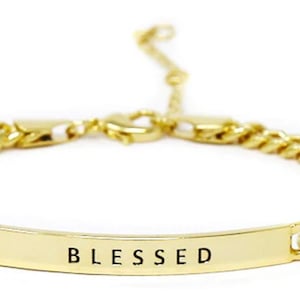 May include: A gold chain bracelet with a gold bar charm that says "BLESSED" in black lettering.