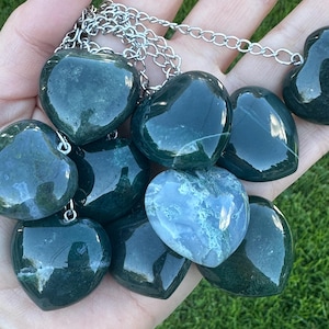 May include: A collection of heart-shaped pendants made of polished green and blue-green stone. Each pendant is strung on a silver-colored chain. The pendants vary in color and pattern, with some having a marbled appearance.