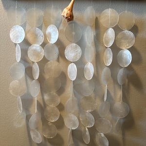 May include: A wall hanging featuring a horizontal piece of driftwood wrapped with twine. Multiple vertical strands of translucent, circular capiz shell discs hang from the wood. A single tan, spiral-shaped seashell is attached to one strand.