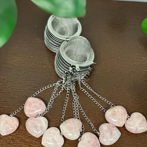 May include: Two stainless steel tea infusers with mesh baskets, each attached to a chain of small links. The chains lead to heart-shaped rose quartz stones. The background is a dark brown surface.