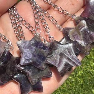 May include: Close-up of star-shaped pendants made of purple and white stone, strung on silver chains. The pendants are polished and have a natural, marbled appearance. The background is a blurred view of green grass.