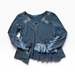 Puede incluir: Una blusa de manga larga en color azul pizarra oscuro con aplicaciones florales de encaje azul claro alrededor del cuello y los puños. La prenda incluye un dobladillo con volantes superpuestos, un panel de tela transparente y arrugada.