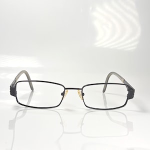 Peut inclure: Lunettes rectangulaires noires avec une fine monture en métal et des verres transparents. Les branches sont grises et présentent un motif texturé près des charnières. Les lunettes sont posées sur un fond blanc.