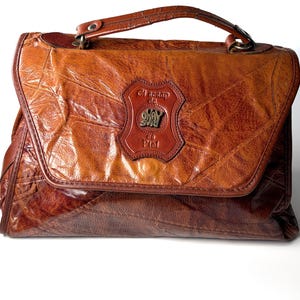 Puede incluir: Un bolso de mano marrón construido con piezas de cuero en patchwork con un acabado brillante. El bolso cuenta con un asa superior y un parche de cuero central grabado con el texto El Scrap da Okay Sac es Piel y un emblema metálico central.