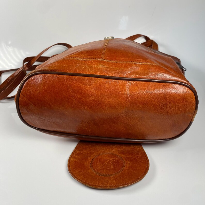 May include: A vintage, brown leather shoulder bag with a rounded shape. The bag features a flap closure and a long, adjustable strap. The leather has a distressed appearance, with visible wear and tear. The bag is a warm, earthy tone.