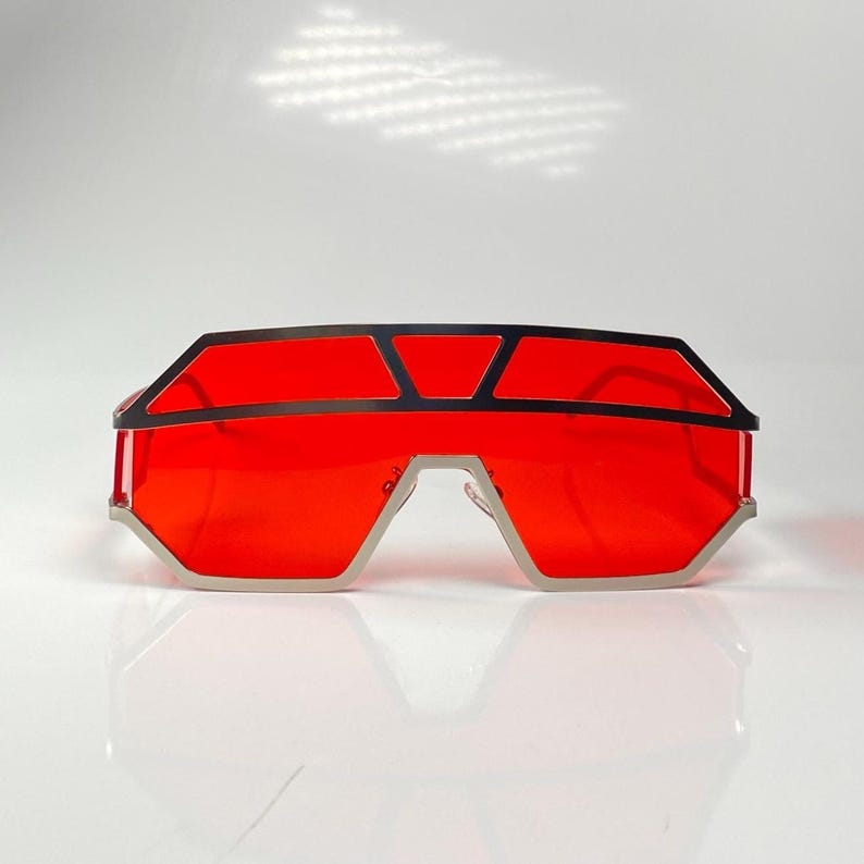 May include: Futuristic sunglasses with a bold, geometric design. The frames are silver with black accents, and the lenses are a vibrant red. The sunglasses have a unique, angular shape, perfect for a statement look.