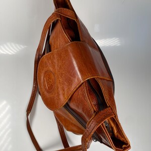 May include: A brown leather backpack with a textured finish. The bag features a front flap with a circular emblem and a zippered pocket. The straps are thin and adjustable. The bag is designed to be worn over one shoulder.