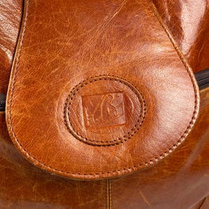 May include: Close-up of a brown leather bag with a flap closure. The flap features a stitched, circular emblem with an embossed design. The leather has a textured appearance and a warm, reddish-brown hue.
