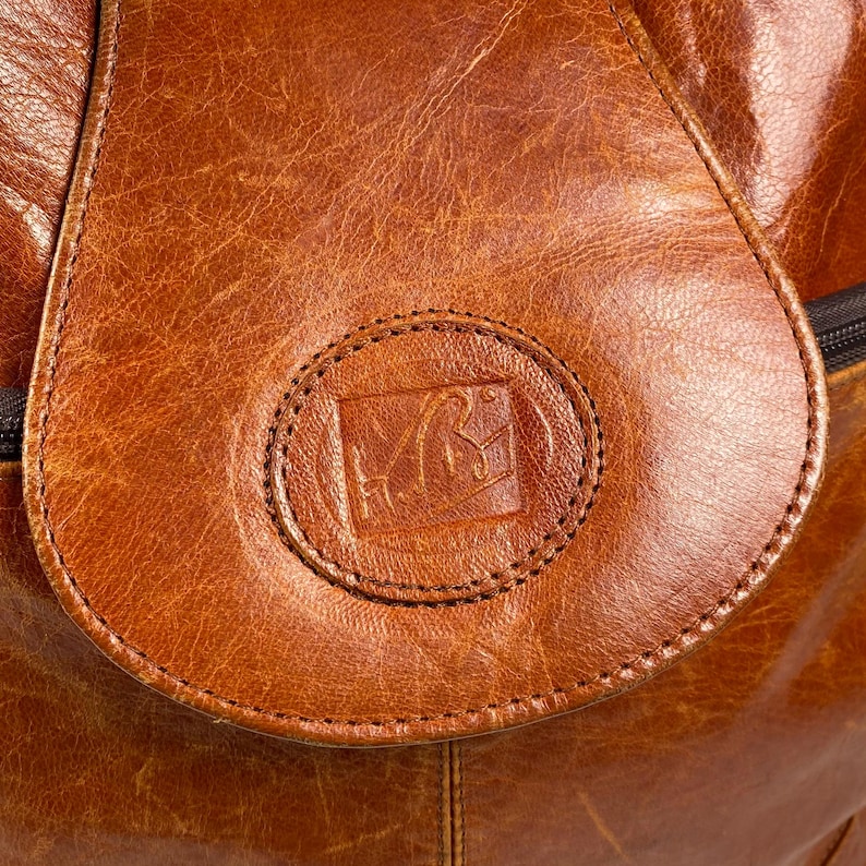 May include: Close-up of a brown leather bag with a flap closure. The flap features a stitched, circular emblem with an embossed design. The leather has a textured appearance and a warm, reddish-brown hue.