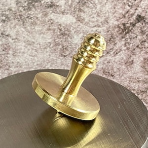 Puede incluir: Una peonza dorada con una parte superior redondeada y estriada y una base circular plana.