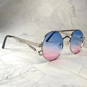 Op de afbeelding: Zilveren ronde zonnebril met een blauwe tot roze verlooplens. De zonnebril heeft een veer scharnier en een metalen frame.