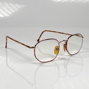 Könnte beinhalten: Ovale Brille mit einem goldfarbenen Metallrahmen und braun-beigen gemusterten Bügeln. Die Gläser sind klar, und die Nasenpads sind gelb. Die Brille ist vor einem weißen Hintergrund platziert.