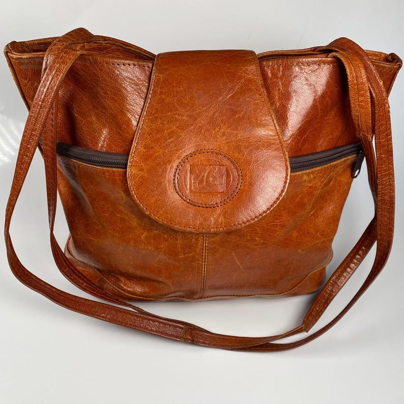 May include: A brown leather shoulder bag with a flap closure and long straps. The bag has a textured surface and a front pocket with a zipper. A circular emblem is embossed on the flap.