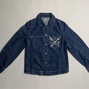 Puede incluir: Chaqueta vaquera azul medio con cierre de botones, cuello y mangas largas. La chaqueta presenta un diseño blanco y gris en el bolsillo delantero y una pequeña etiqueta roja. Chaqueta vaquera clásica.