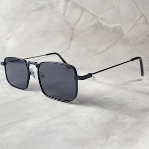 Puede incluir: Gafas de sol cuadradas negras con lentes oscuras. Las gafas tienen una montura fina de metal negro y patillas rectas. Las lentes son de color gris oscuro y el diseño general es moderno y minimalista.