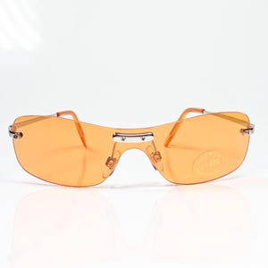 May include: Rimless sunglasses featuring a single continuous orange lens and silver-tone metal hardware. The bridge and hinges are secured with small screws. A circular sticker on the right lens displays the text UV400, indicating protection.
