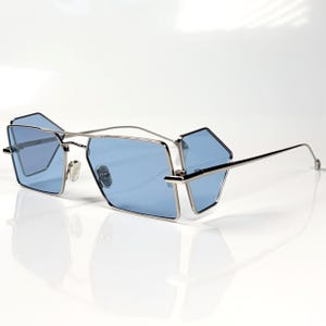 Puede incluir: Gafas de sol con montura de metal plateado y lentes tintadas en azul claro. Las lentes presentan una forma geométrica multifacética distintiva con un diseño dividido. Las varillas de metal fino se extienden desde los lados y el puente conecta las dos secciones.
