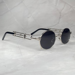 Puede incluir: Gafas de sol redondas con lentes negras y montura de metal plateado. La montura presenta un diseño único con puente y detalles laterales. Las patillas son negras. Las gafas están sobre un fondo claro.