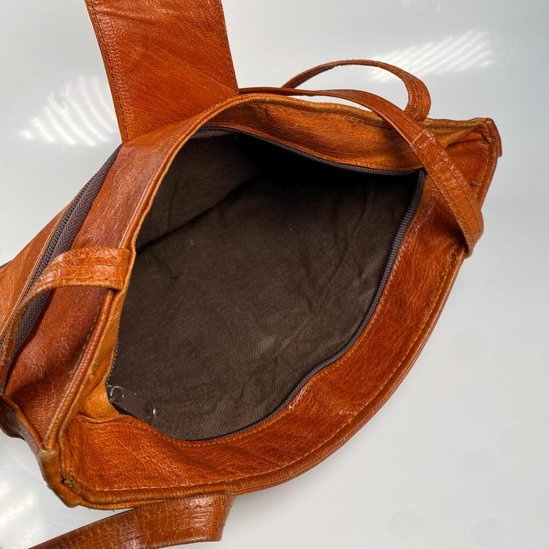 May include: Open view of a brown leather tote bag. The bag has a zipper closure and a brown interior. The bag has two handles and a shoulder strap. The leather has a textured appearance.