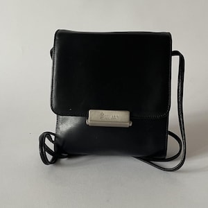 Può includere: Borsa a tracolla in pelle nera con chiusura a patta e chiusura in metallo argentato. La borsa ha una tracolla lunga e sottile. Il marchio "ASSIMA" è visibile sulla chiusura. La borsa è di forma quadrata.