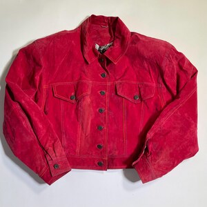 May include: A bright red suede jacket with a classic button-up design. The jacket features two chest pockets, buttoned cuffs, and a collared neckline. The stitching is a contrasting orange color.