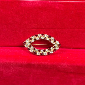 Op de afbeelding: Een ovaalvormige broche met een goudkleurig metalen frame, bezet met afwisselend heldere en dieprode gefacetteerde strass-steentjes. Het ontwerp creëert een hol midden, waarbij de stenen in twee rijen een marquise-achtige vorm vormen.