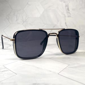 May include: Black square-shaped sunglasses with a gold metal frame and black lenses.