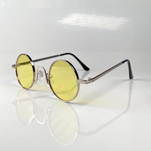 May include: Round sunglasses featuring bright yellow lenses and a thin, polished silver-tone metal frame. The eyewear includes adjustable nose pads and slender temples with black plastic tips, providing a clear view of the circular lens design.
