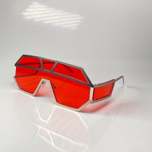 May include: Futuristic red sunglasses with a silver metal frame. The lenses are a vibrant red, and the frame has a geometric design. The sunglasses have a unique, angular shape, and the temples are thin and silver.