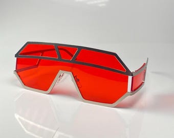 Peut inclure: Lunettes de soleil futuristes avec une monture argentée et des verres rouge vif. Le design géométrique présente une forme angulaire audacieuse, offrant une esthétique moderne. Les verres sont d'un rouge vif uni, contrastant avec la monture argentée.
