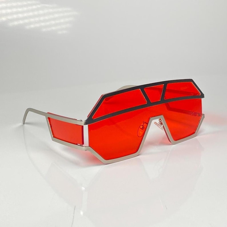 May include: Futuristic-style sunglasses with a bold design. The sunglasses feature a silver frame and vibrant red lenses. The top of the frame has a black accent, adding contrast to the overall look. The glasses have a unique, angular shape.