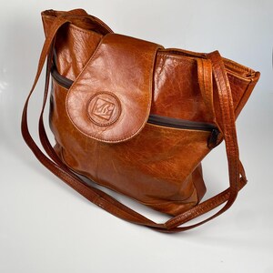 May include: A brown leather tote bag with a flap closure and a long shoulder strap. The bag features a zipper pocket and a circular emblem on the flap. The leather has a distressed appearance.