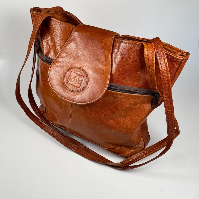 May include: A brown leather tote bag with a flap closure and a long shoulder strap. The bag features a zipper pocket and a circular emblem on the flap. The leather has a distressed appearance.