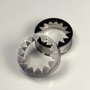 May include: Two silver ear gauges with a starburst design. One gauge has a black center and the other has a silver center.
