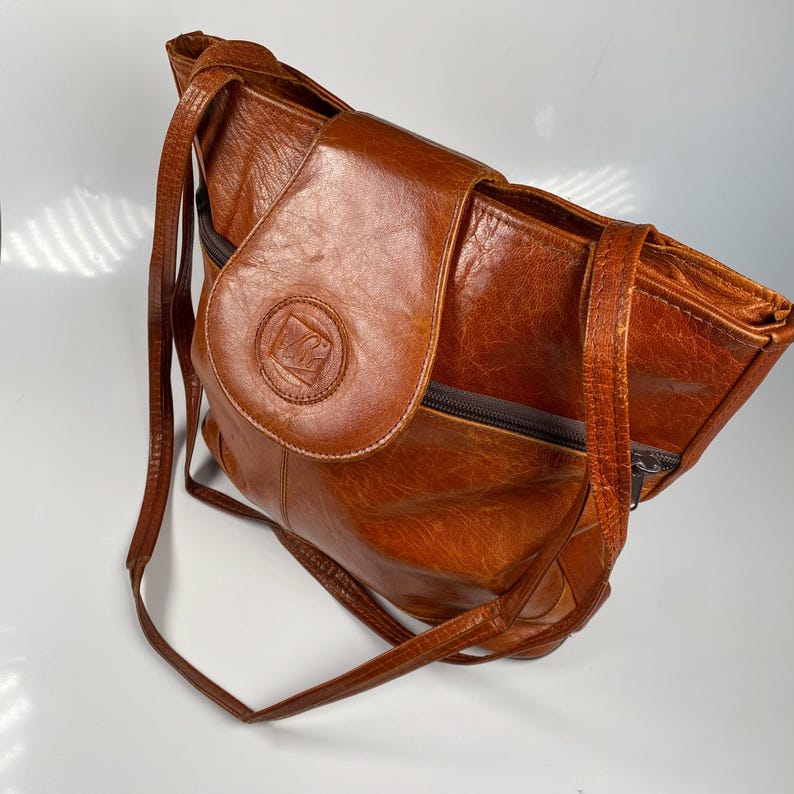 May include: A brown leather shoulder bag with a flap closure and long strap. The bag has a circular emblem on the flap and a zipper closure. The leather has a distressed appearance.