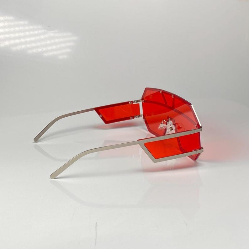 May include: Red geometric sunglasses with silver-colored frames. The lenses are a vibrant red, and the frames have a modern, angular design. The sunglasses are set against a white background.