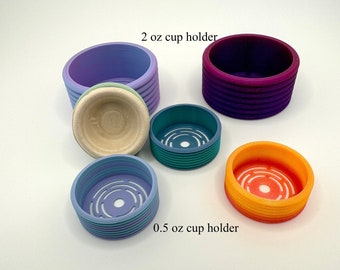 May include: Set of five 3D printed cup holders in various colors, including purple, teal, and orange. The cup holders are designed to hold 0.5 oz and 2 oz cups. The text "2 oz cup holder" and "0.5 oz cup holder" is printed below the cup holders.