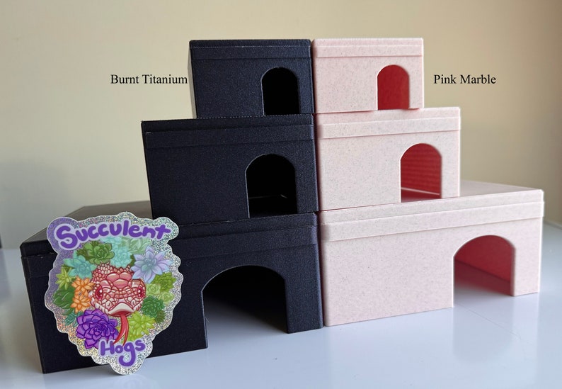 Op de afbeelding: Een set van drie gestapelde hamsterhuisjes, &eacute;&eacute;n in gebrand titanium en twee in roze marmer. De huisjes hebben gebogen openingen en zijn gemaakt van een getextureerd materiaal. Een sticker met de tekst "Succulent Hogs" zit op het onderste huisje.