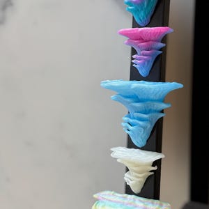 May include: A vertical display of colorful, layered mushroom sculptures. The mushrooms are in shades of blue, pink, white, and iridescent colors, attached to a black vertical support. The background is a neutral white.