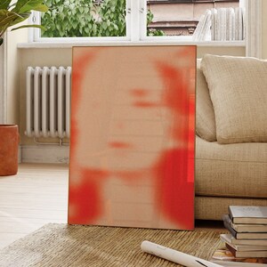 May include: A large, abstract painting with a blurred, pink and red design hangs on a wall in a living room. The painting is leaning against a white radiator and is in front of a beige couch.