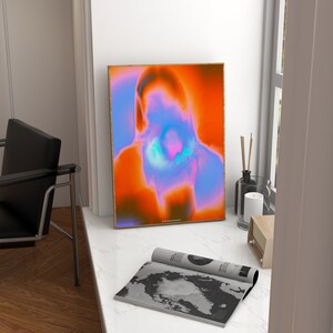 May include: A framed abstract art print with a blurred image of a person's face in shades of orange, blue, and pink. The print is in a gold frame and is hanging on a white wall.