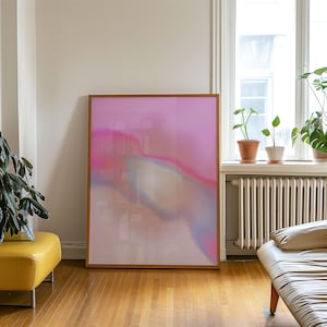 May include: A large framed artwork with an abstract design in shades of pink, purple, and white. The artwork is leaning against a wall in a room with hardwood floors and a yellow ottoman. The frame is a warm wood tone.