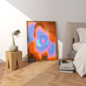 May include: A framed abstract art print with a blurred image of a person's face in shades of orange, pink, and blue. The print is hanging on a white wall in a room with a wooden floor.