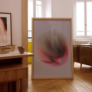 May include: A large framed artwork with an abstract design. The artwork features a central blurred shape in red and pink against a light grey background. The frame is light-coloured wood. The artwork is on a wooden floor.