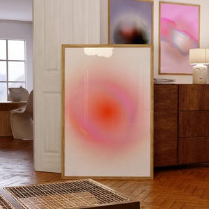 May include: Aesthetic wall art featuring abstract designs in a room setting. The large framed artwork has a gradient of pink and orange hues. Two additional framed pieces display blurred, colorful compositions. The room has wooden floors and furniture.